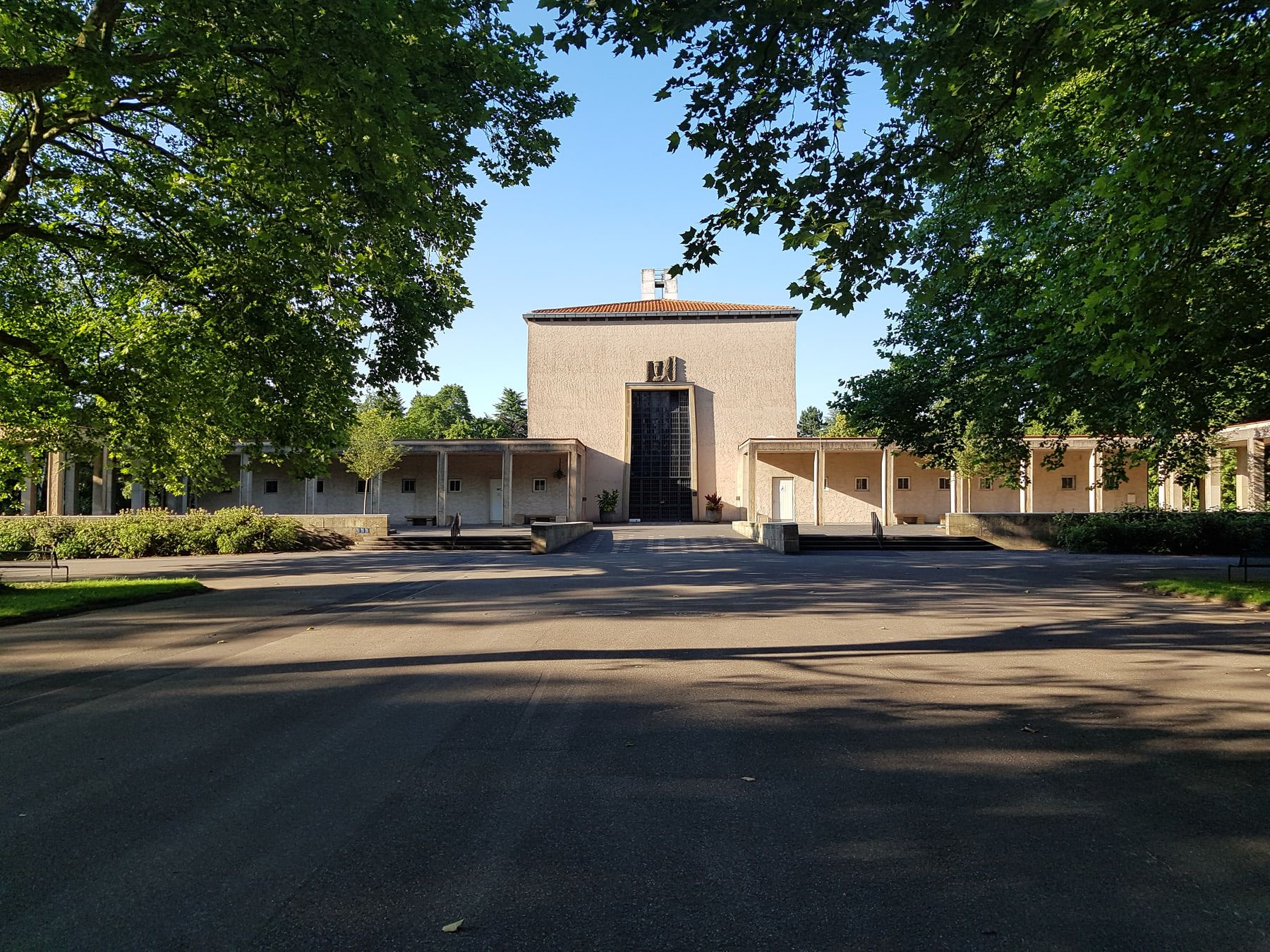 Trauerhalle mit vorgelagertem Platz und seitlichen Kolonnaden unter altem Baumbestand auf Westfriedhof Köln