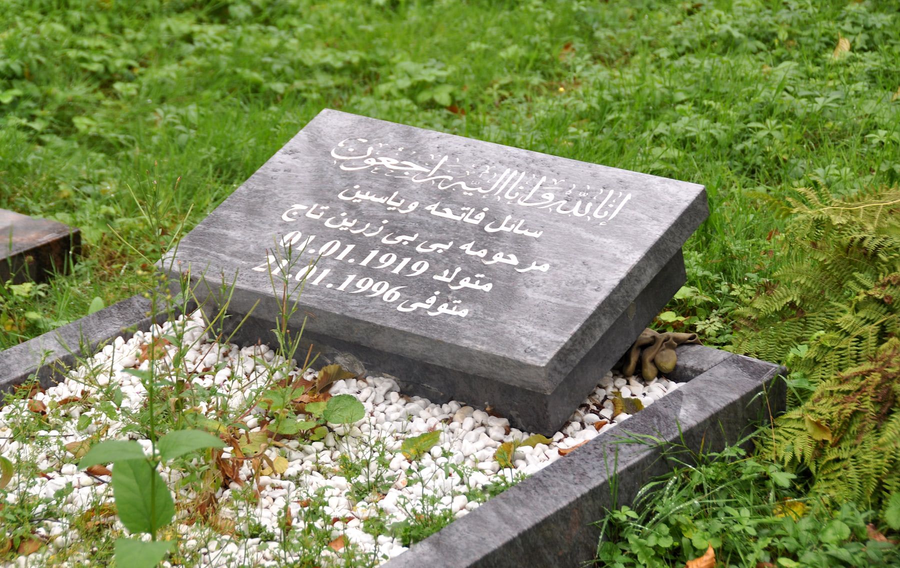 Dunkler Grabstein mit weißer Inschrift in arabischer Schrift in einem muslimischen Grabfeld, eingefasst von weißen Kieseln und Farnen auf Westfriedhof Köln